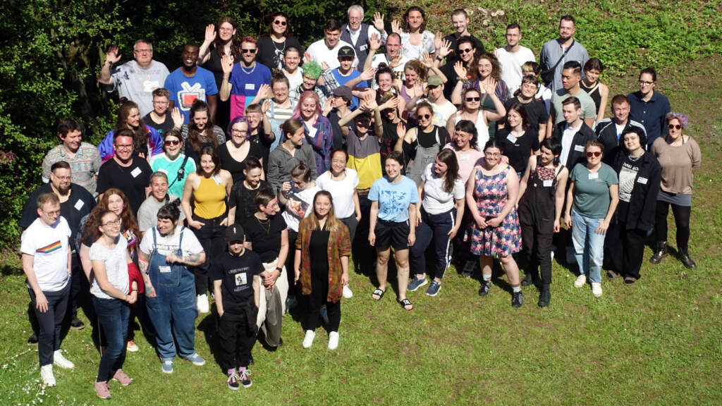 Gruppenfoto der Teilnehmenden der Bundesvernetzung 2023 auf einer Wiese bei Sonnenschein. Es wurde von schräg oben aufgenommen.