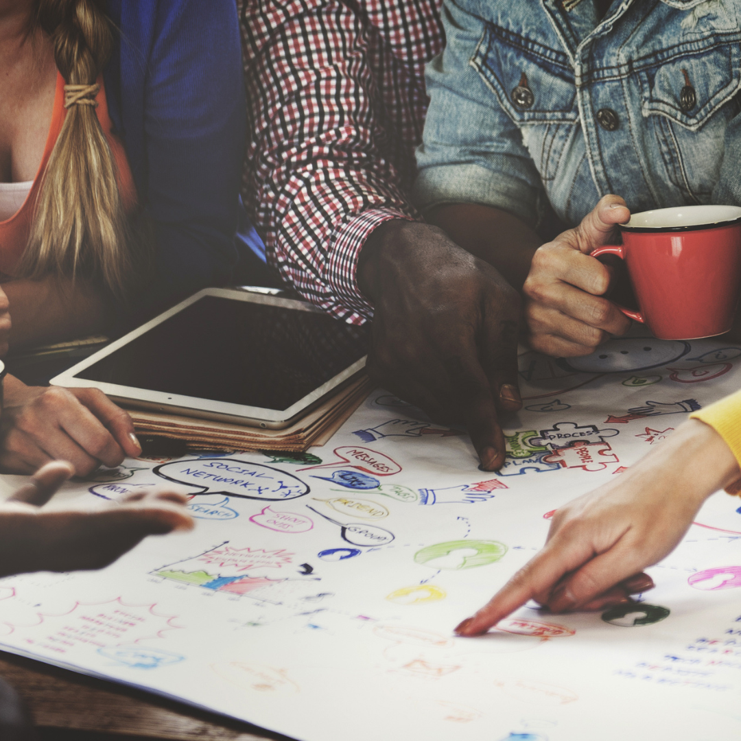 Ein bunt beschriftetes Flipchart-Papier liegt auf einem Tisch. Mehrere Menschen sitzen darum herum und zeigen auf Dinge auf dem Papier.