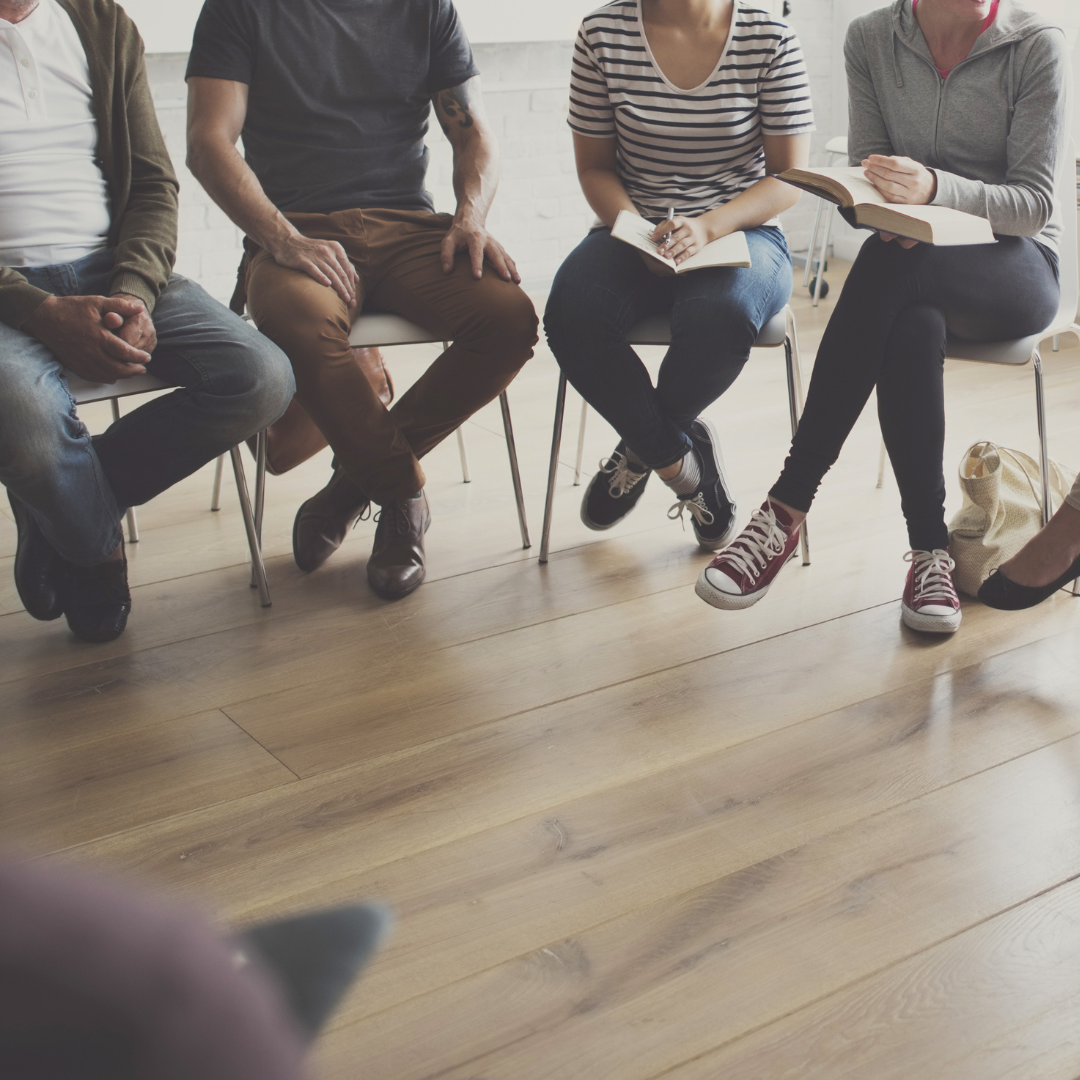 Beine und Oberkörper von vier Personen, die in einem Stuhlkreis sitzen. Eine Person hält ein Buch, eine Person macht sich Notizen.